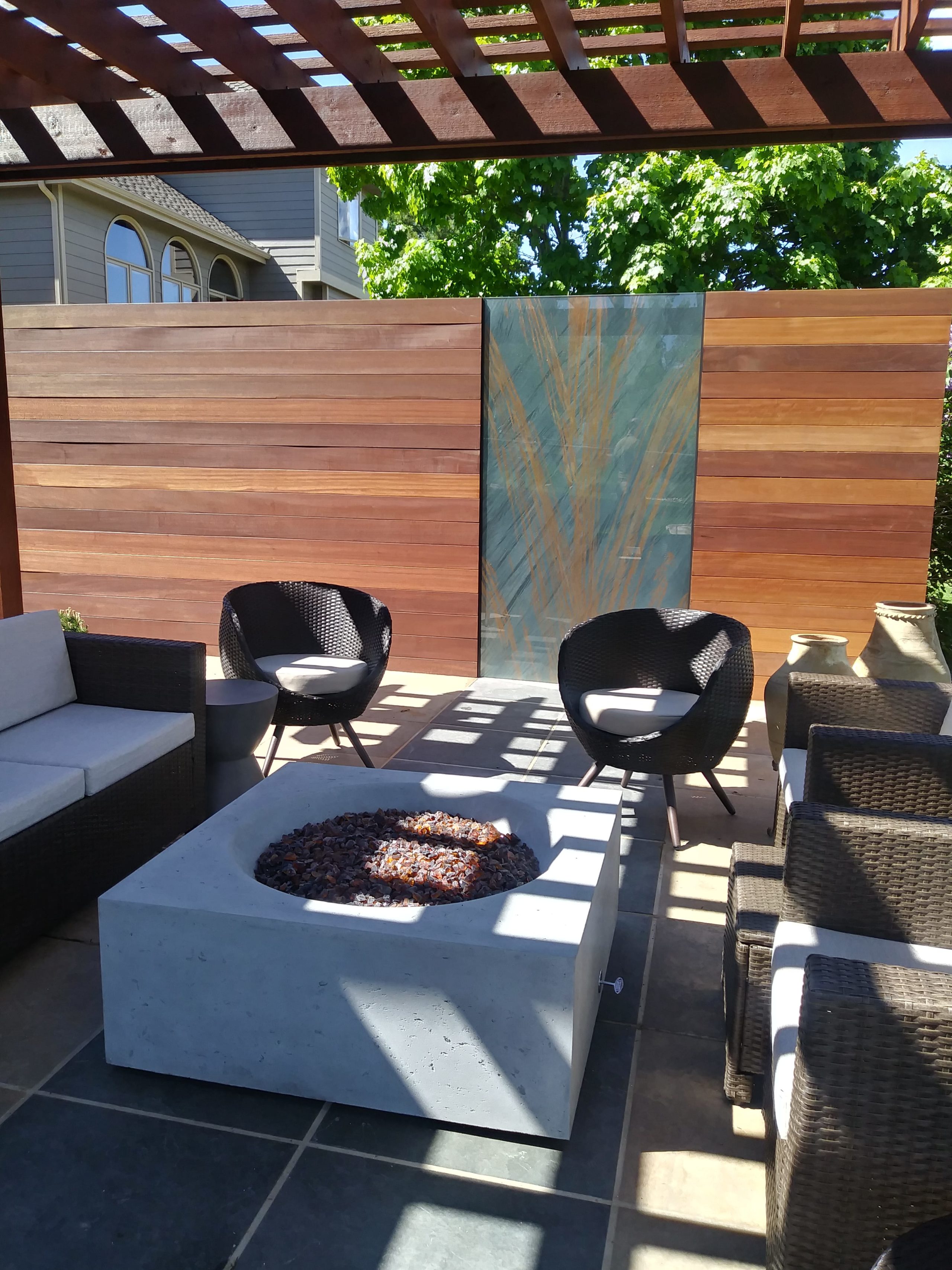 Large pergola over top of a tile patio with seating area around a concrete bowl fire pit.