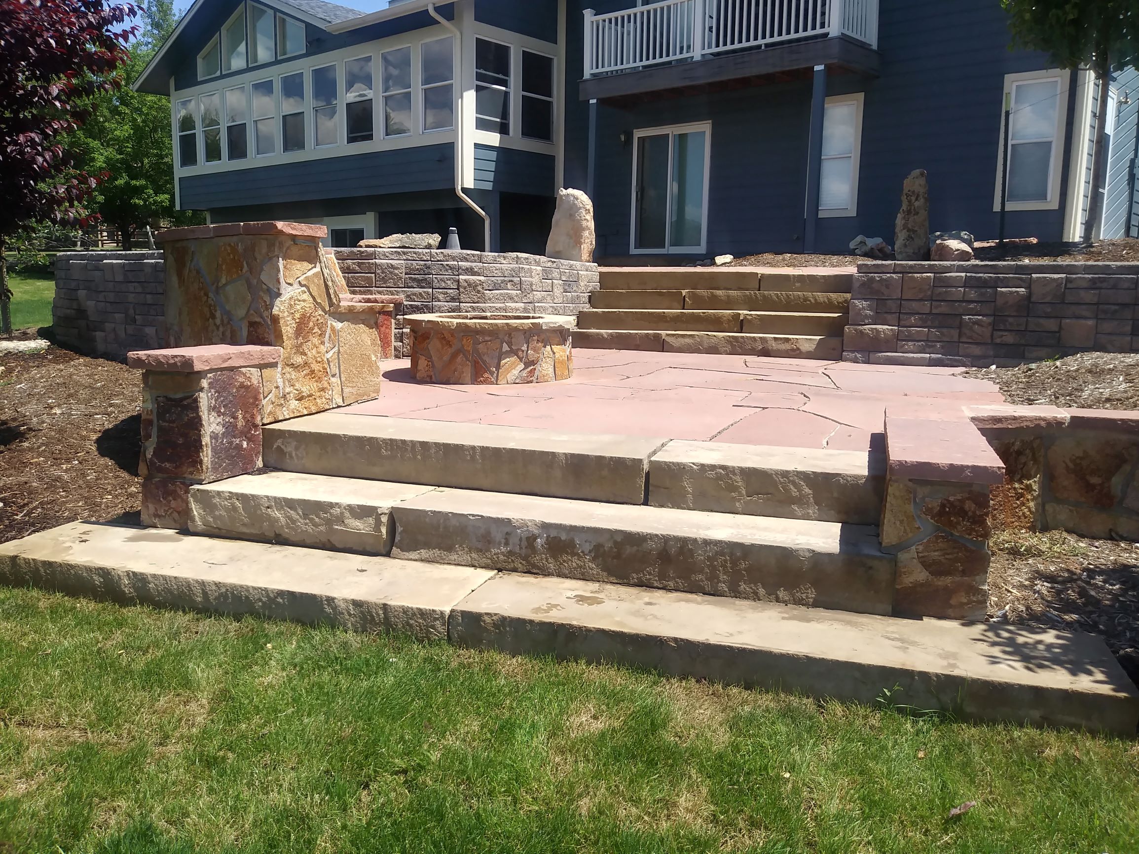 Flat stone steps with flat stone patio and fire pit. Brick edged raised beds topped with mulch.