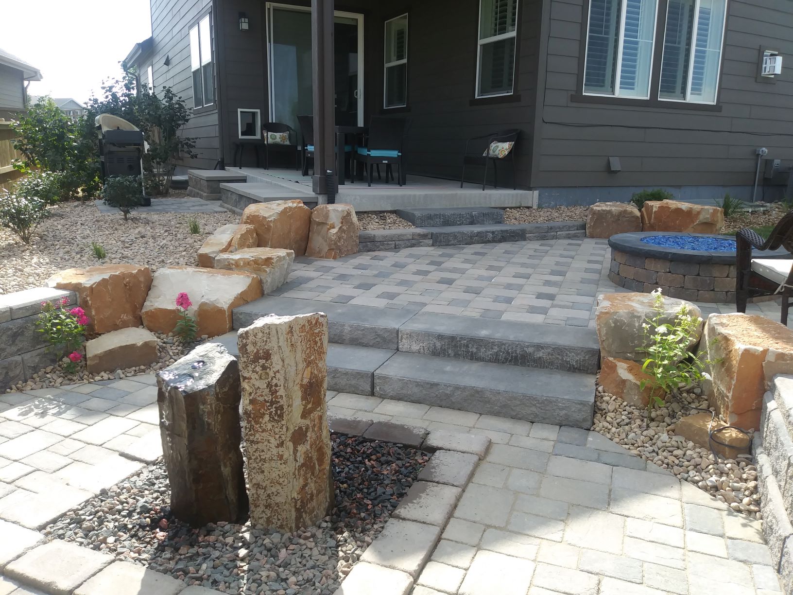 Brick laid patio with water feature and flat stone steps bordered by small boulders.