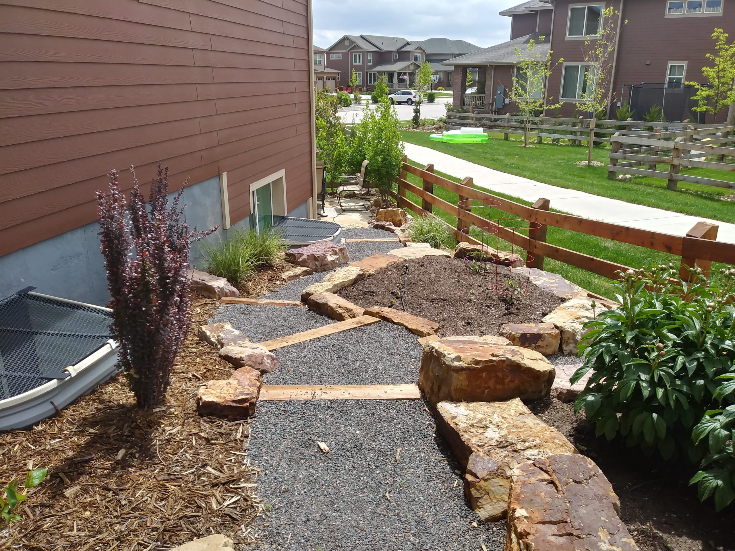 Gravel path with wood steps. Edged with large rocks and mulch beds with plants and bushes.