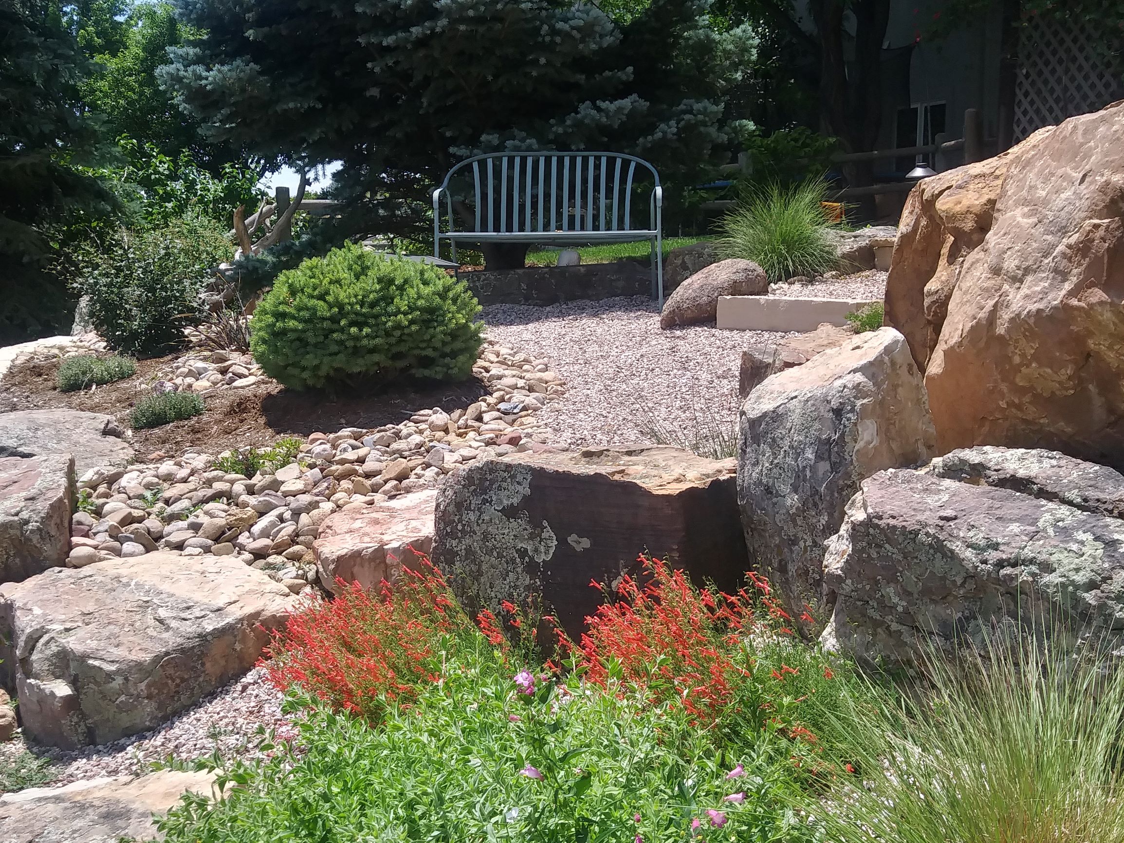 Gravel path leading to a bench. Bordered by large decorative rocks as well as bushes and plants.