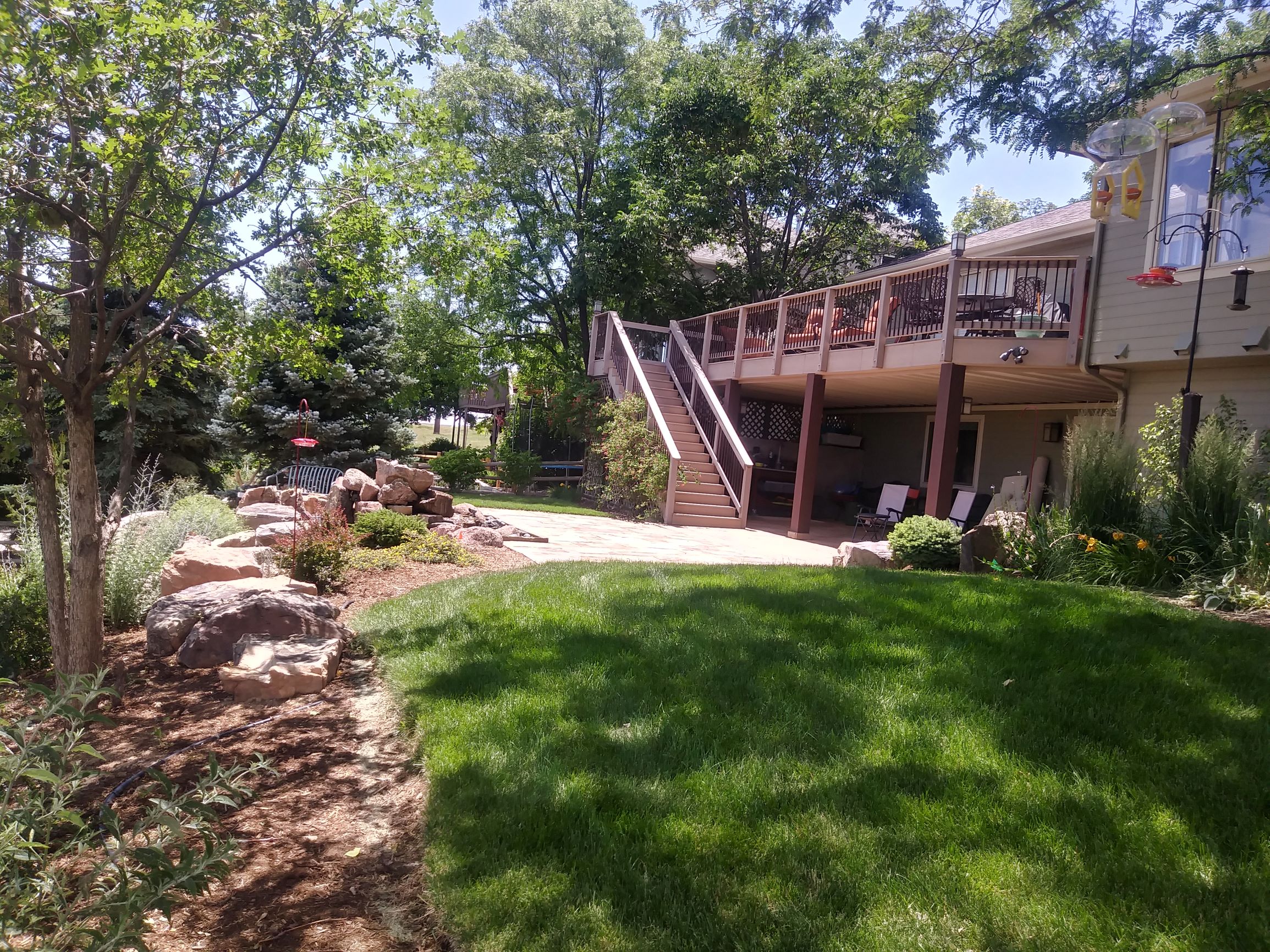 Second story deck leading to flat stone patio, bordered by large rocks, green grass, and many trees and bushes.