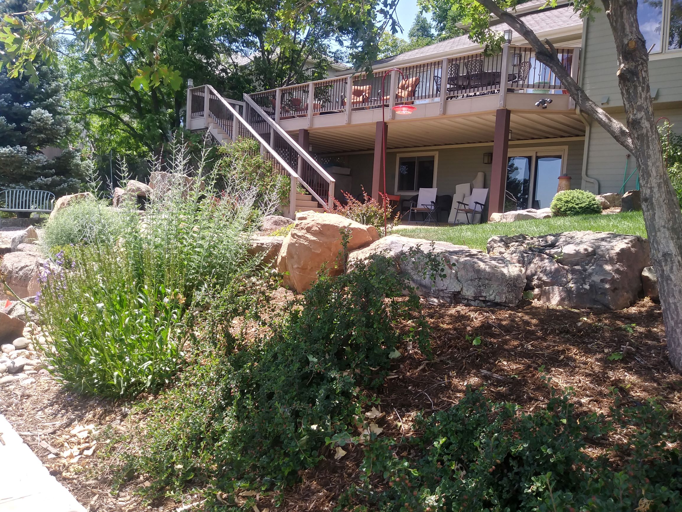 Second story deck with adjacent large rocks and plenty of bushes and plants.