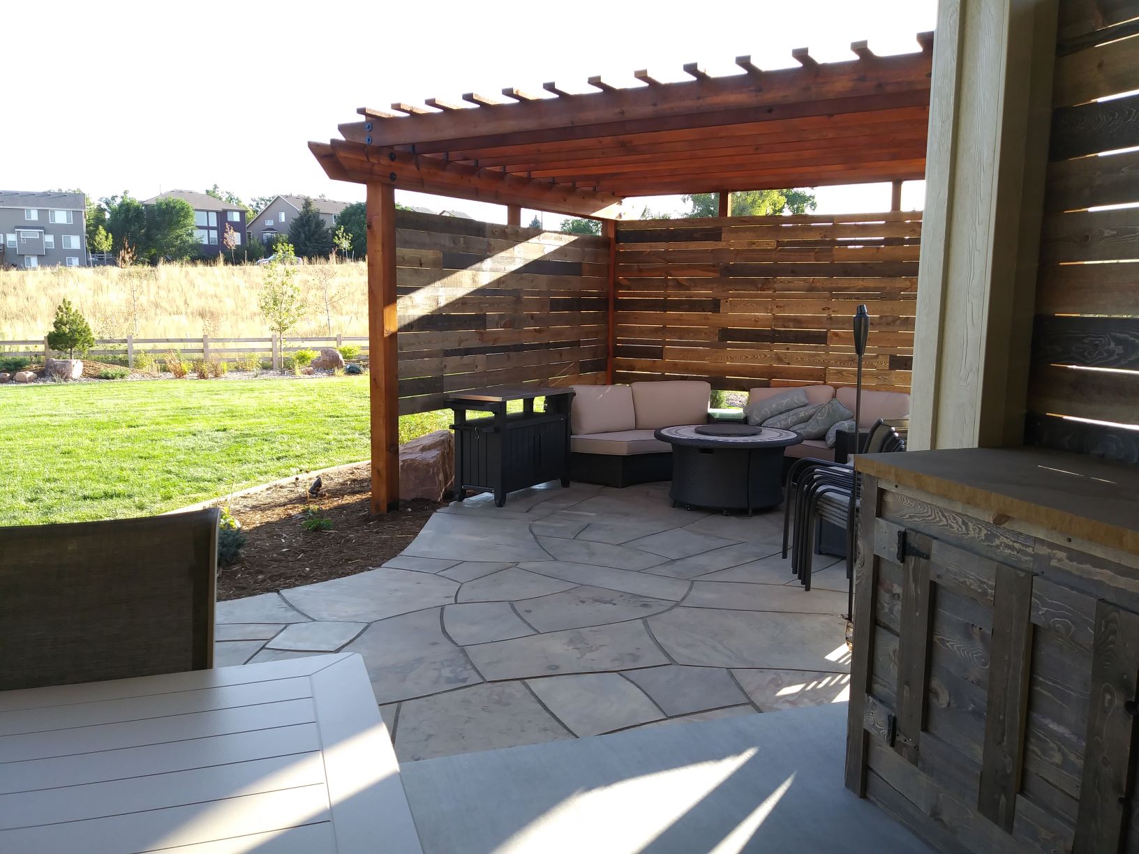 Pergola over flat stone patio with wall for privacy and wind control.