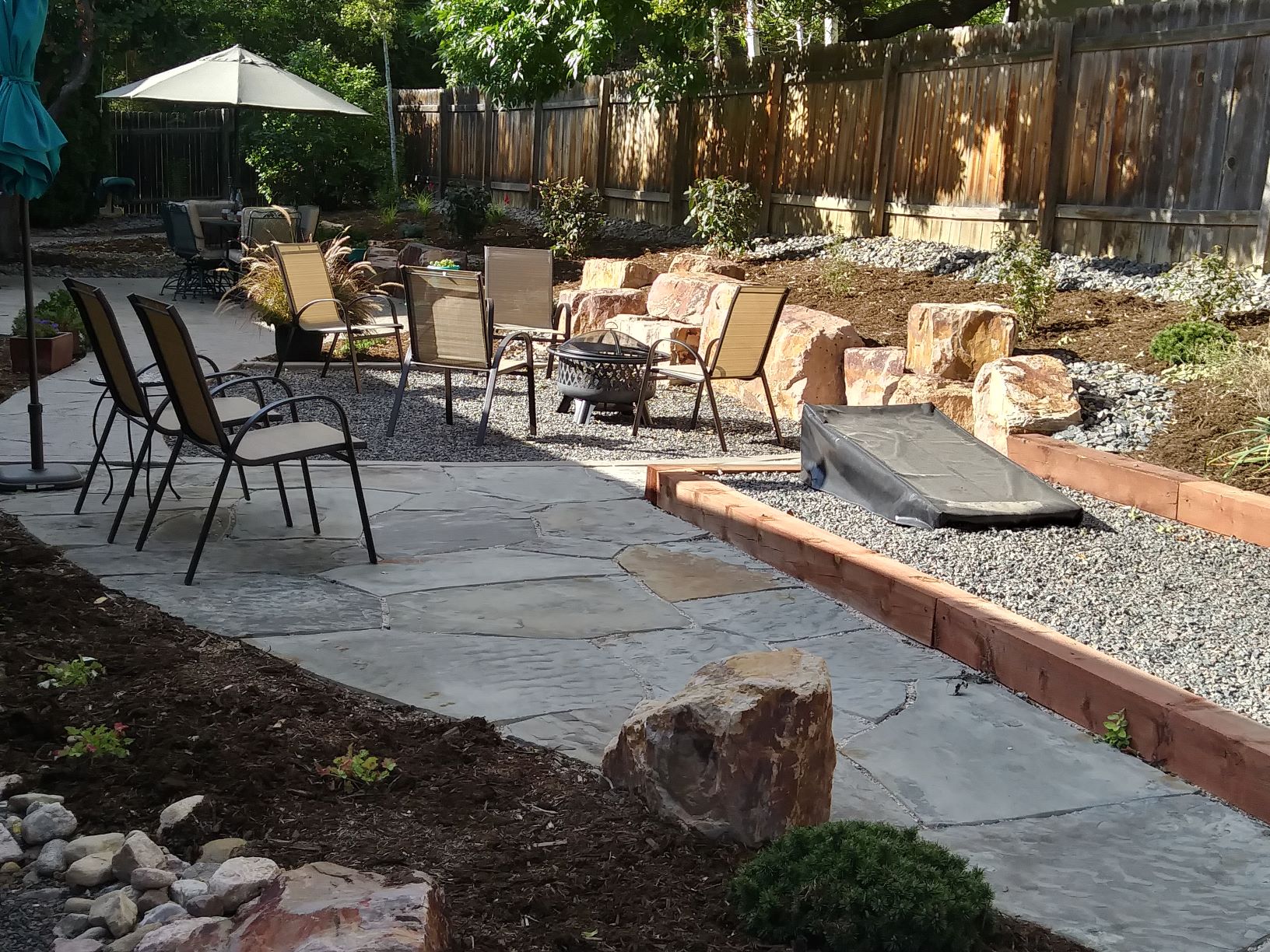 Backyard seating area with flat stone patio and gravel chair area. Surrounded by mulch with low water bushes and plants.