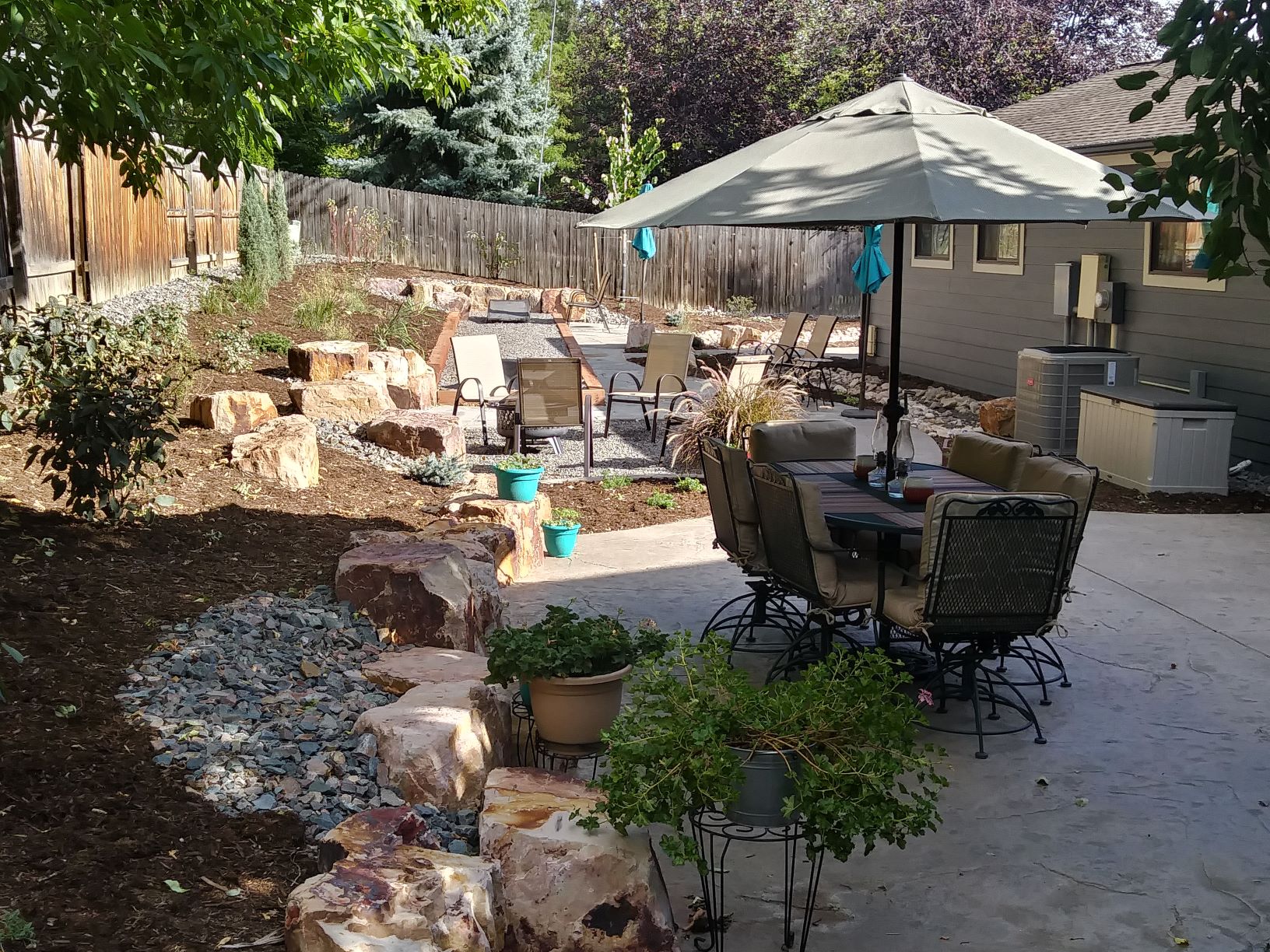 Backyard seating area with flat stone patio and gravel chair area. Surrounded by mulch with low water bushes and plants.
