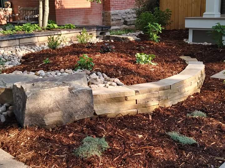 Brick edged raised flower bed with rocks and mulch. Filled with beautiful plants and bushes.