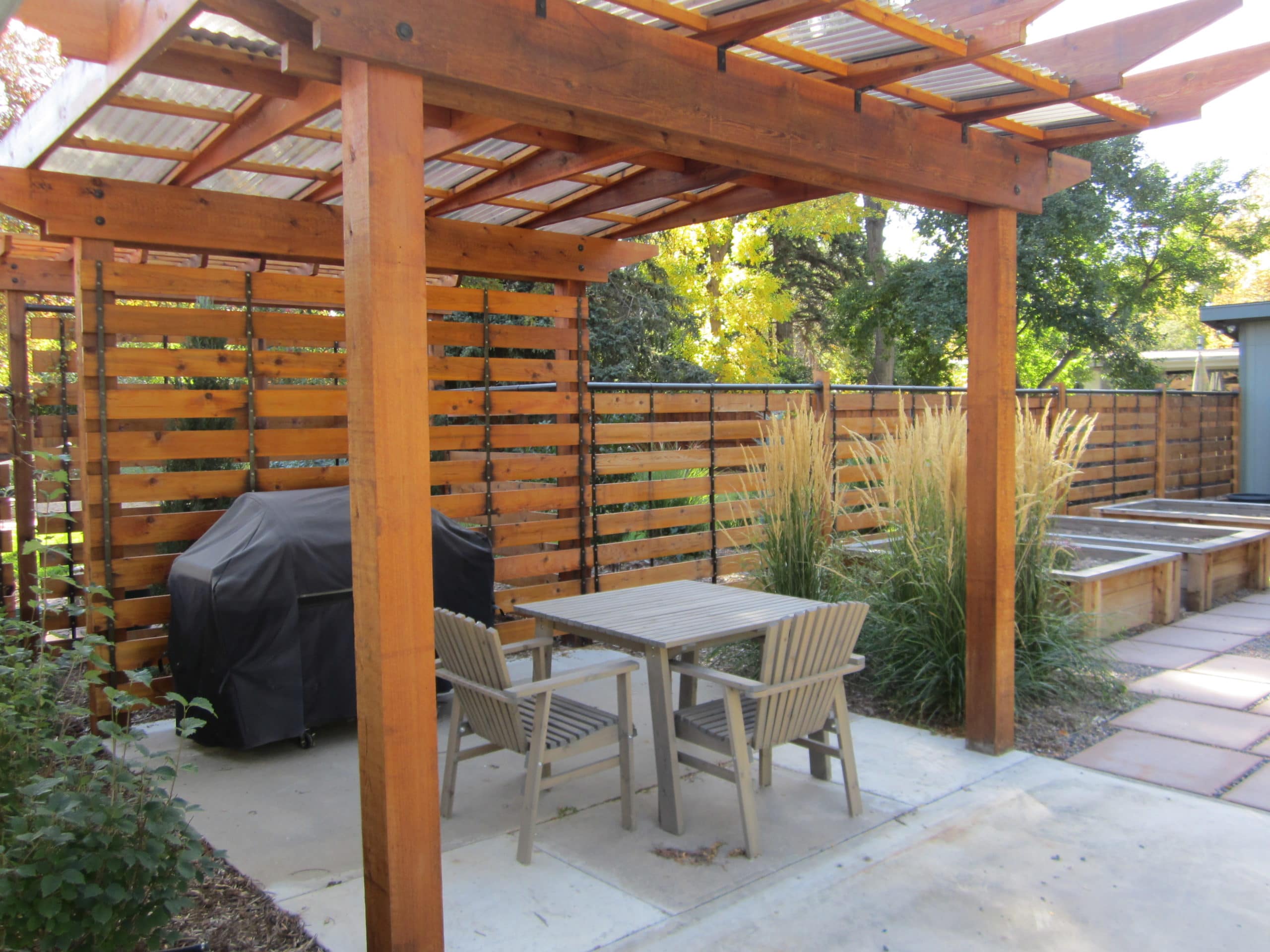 Pergola with dining area. stepping stone path to raised garden beds surrounded by pea gravel and mulch with bushes and plants.