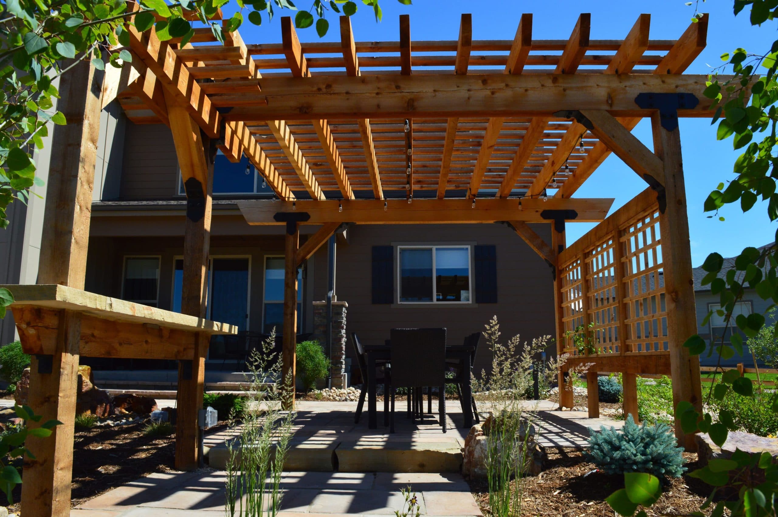 Pergola with dining area on top of brick laid patio surrounded by mulch and rocks with bushes and plants.