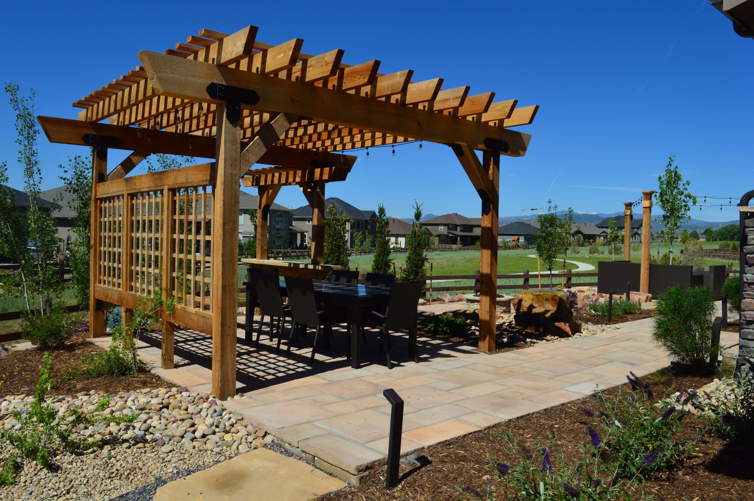 Pergola with dining area on top of brick laid patio surrounded by mulch and rocks with bushes and plants.