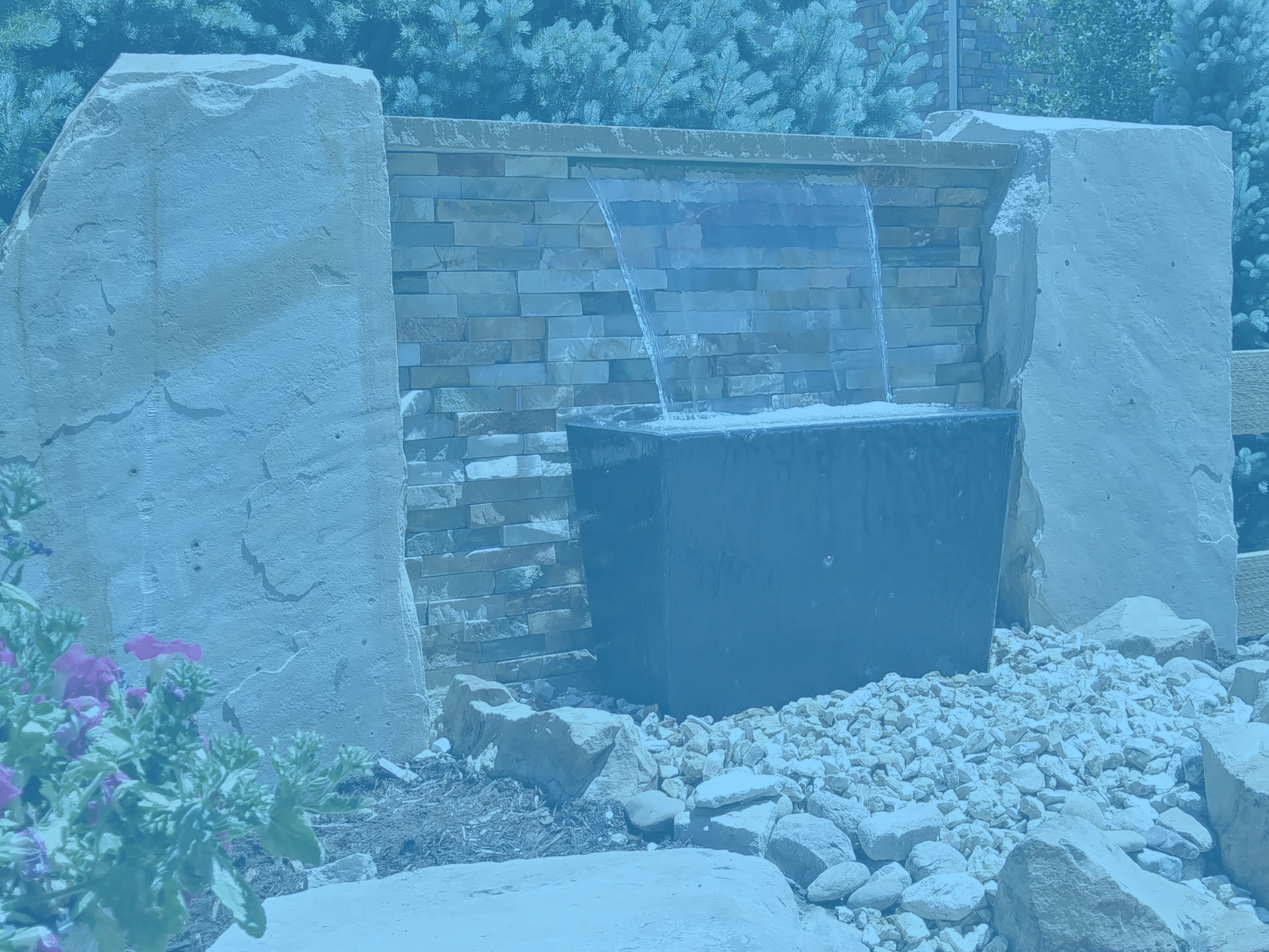 Small water feature surrounded by large decorative rocks, landscape rocks, and mulch with plants and bushes.